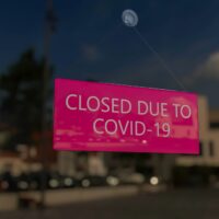 Close-up on a closed sign in the window of a shop displaying the message "Closed due to Covid-19". The glass reflects the day city