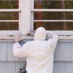 A house painter in a hazmat suit scrapes off dangerous lead paint from a window sill.